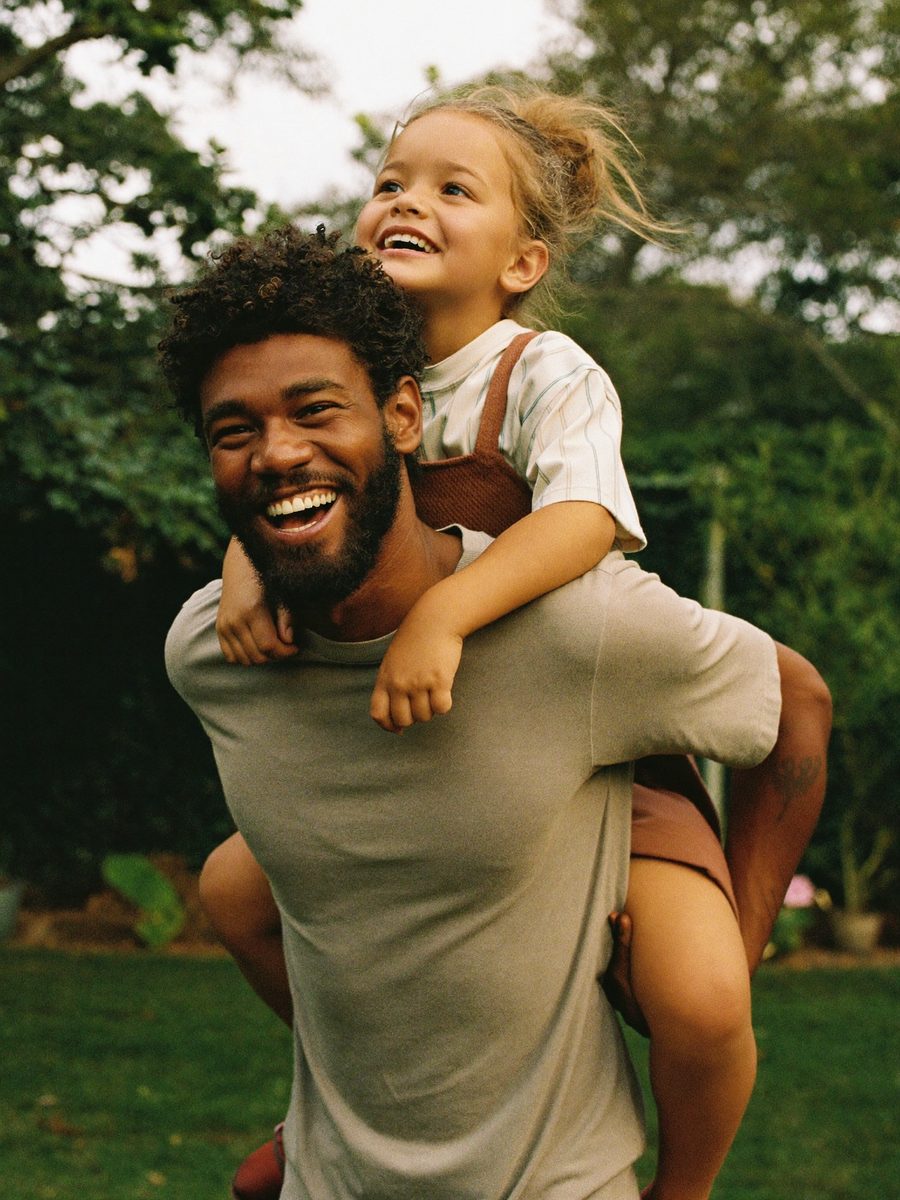 A father laughing with his young daughter on his back in a sunny garden