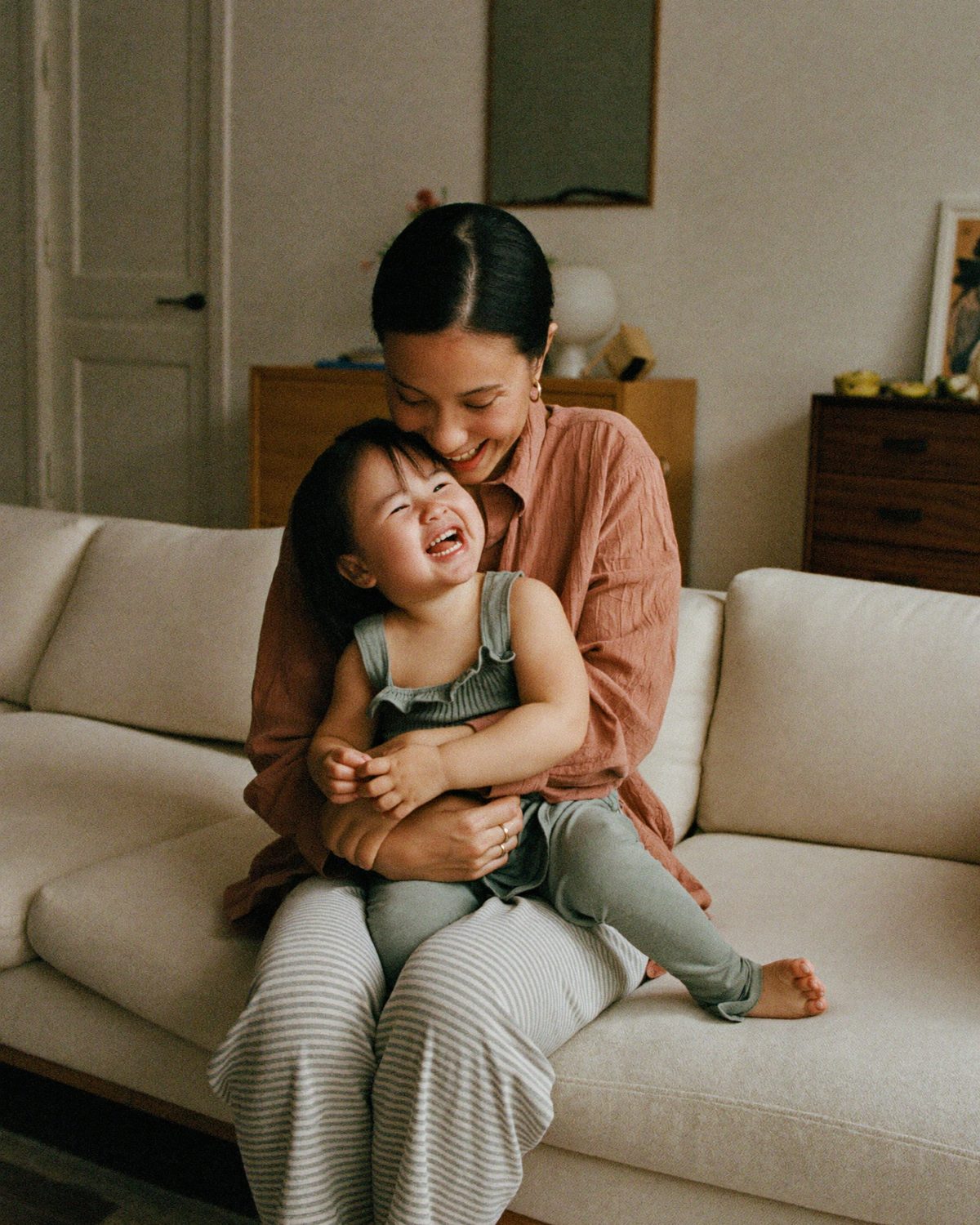 A mother laughing with her toddler daughter on her lap on a cream sofa in soft morning light