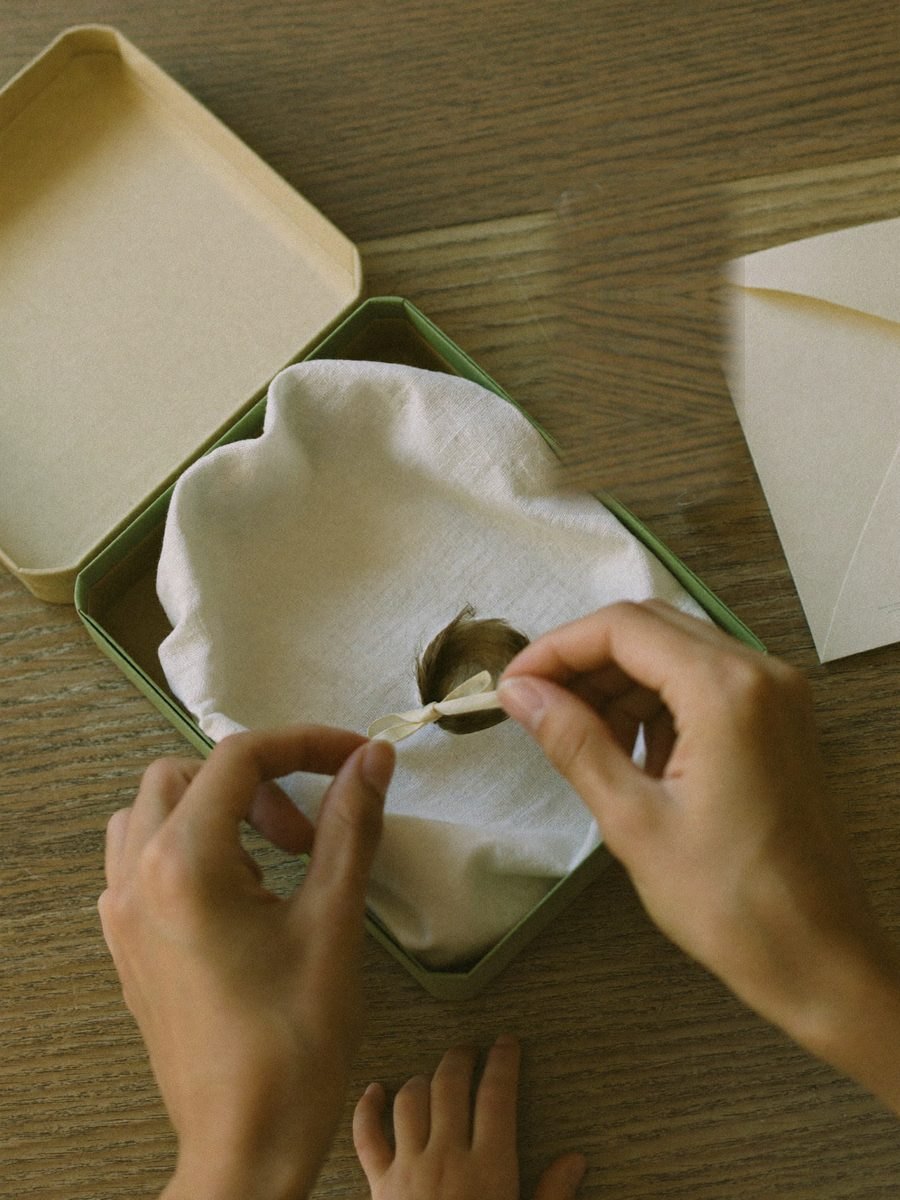 A mother's hands placing a small lock of hair tied with ribbon into a linen-lined keepsake box before it is sealed, a child's hand reaching in