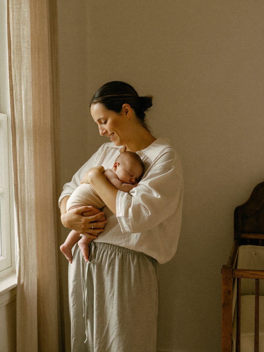 A mother holding her sleeping newborn by a nursery window